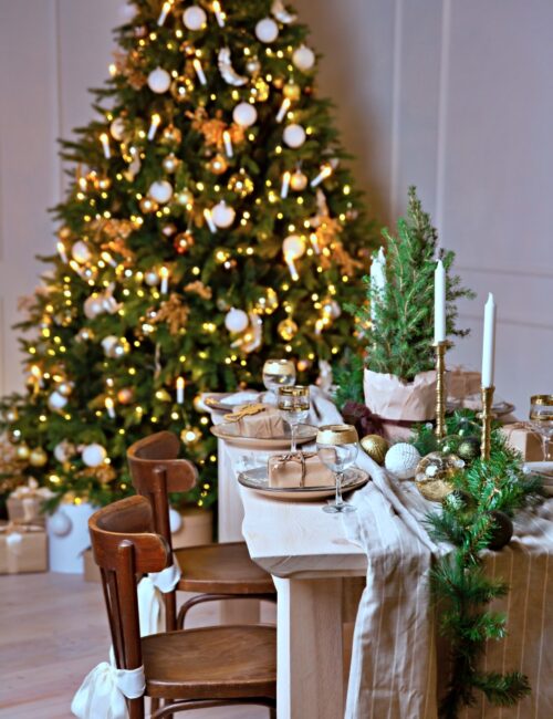 Christmas or New Year festive table setting. The festive wooden dining table is decorated with spruce garland and white candles. Christmas gift on a plate.