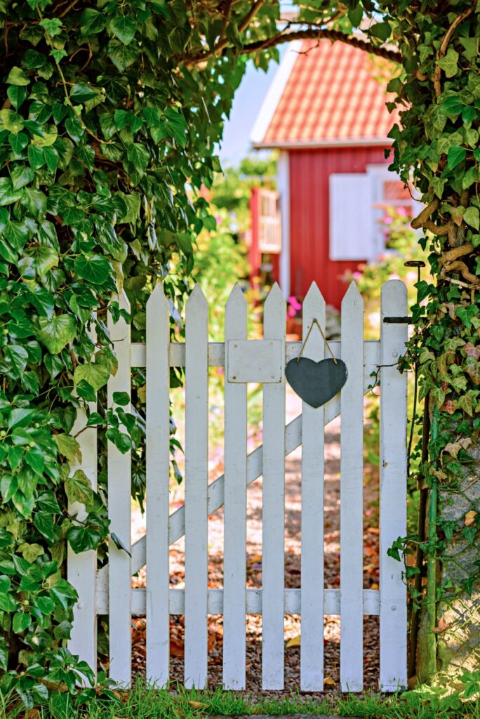 White wooden garden gate with heart and number plate (number removed). Red wooden cabin blurred in background, Hedgerows beside gate.
