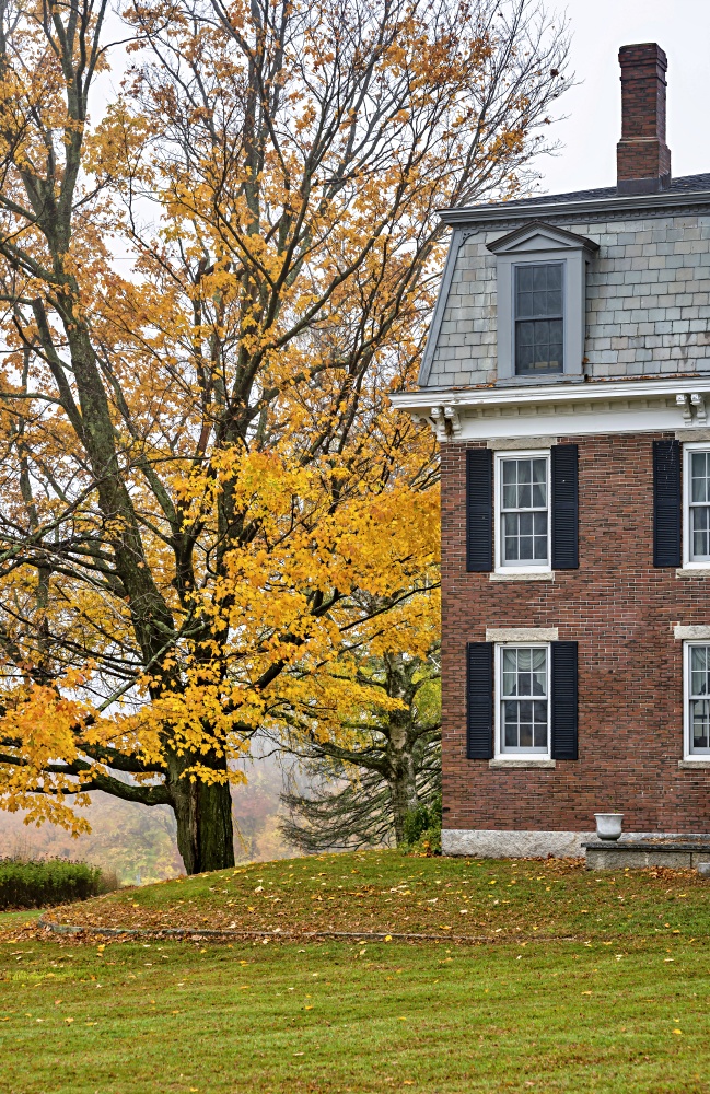 Landscape with a traditional Victorian style old brick house with an attic and a chimney and autumn orange maple tree on the hillside in foggy weather in Vermont